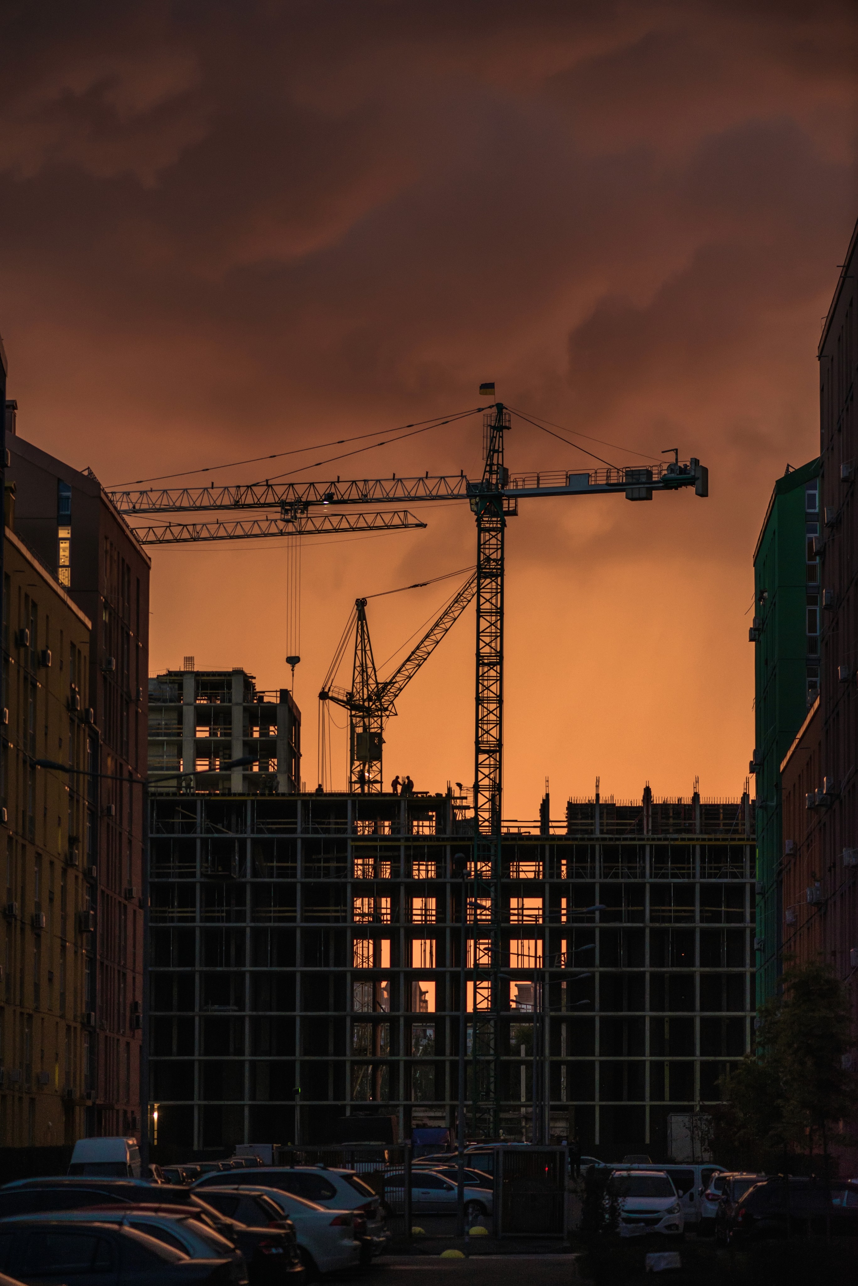 Tower crane silhouettes at an urban building construction site during a dramatic orange sunset.